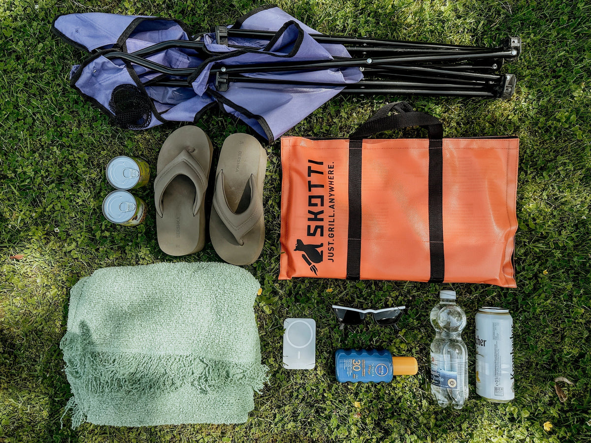 Auf dem bild sind folgende Gegenstände abgebildet: lila Campingstuhl, Dosen, Schuhe, grüne Decke, Powerbank, Sonnenbrille, Sonnencreme, Wasser, Getränkedose, Skotti
